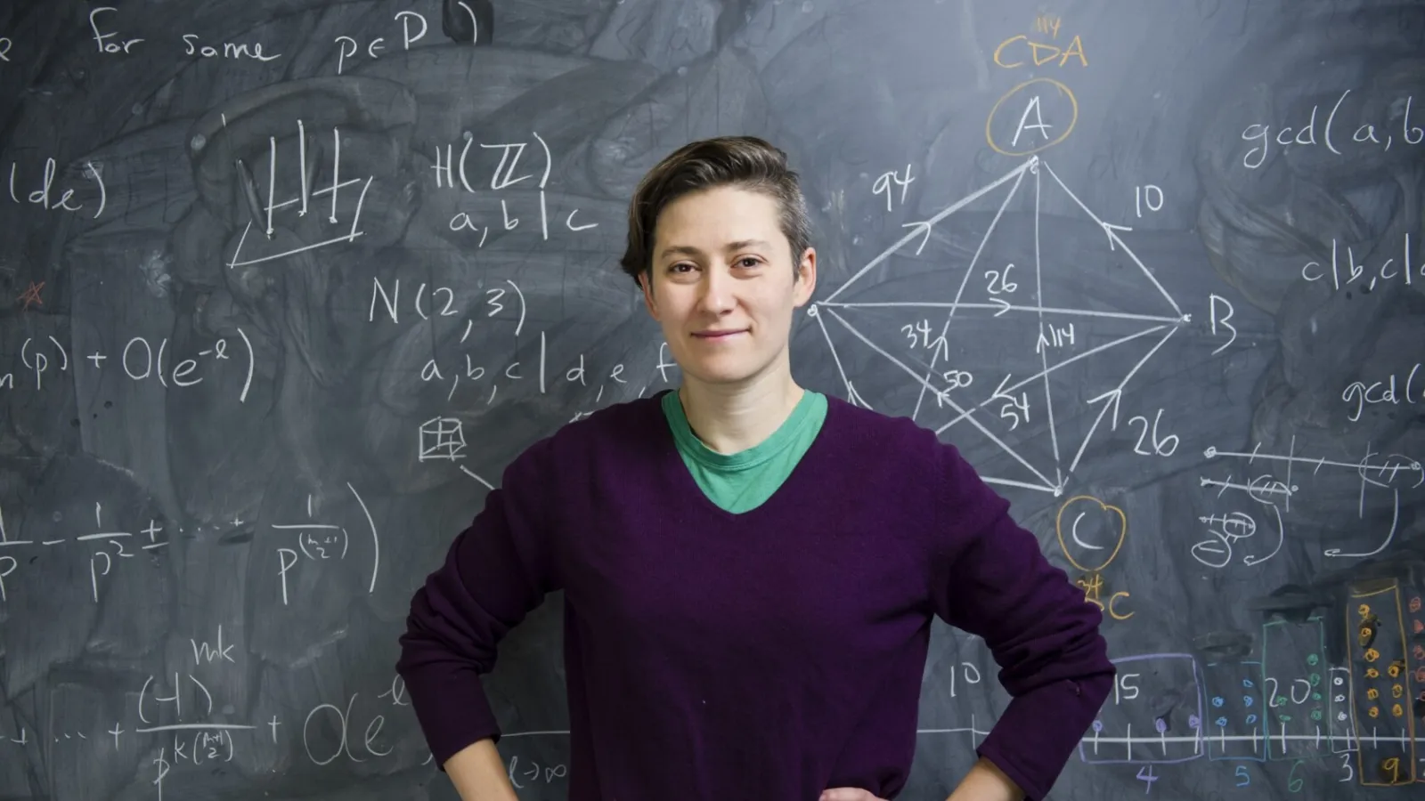 Moon Duchin standing in front of a chalkboard with equations written on the background. 