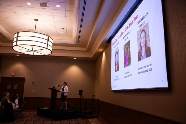 Elly Kaizar standing at a podium with a screen presentation of the award winners